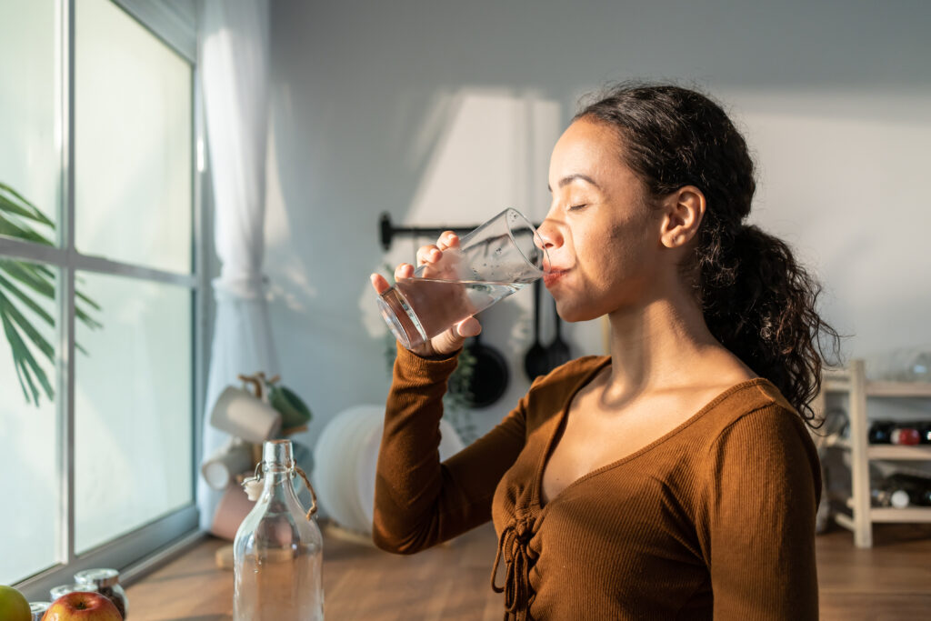Young beautiful Latino woman holding clean water into glass in kitchen. Attractive active thirsty girl drink or take a sips of mineral natural in cup for health care and wellbeing in kitchen in house.
