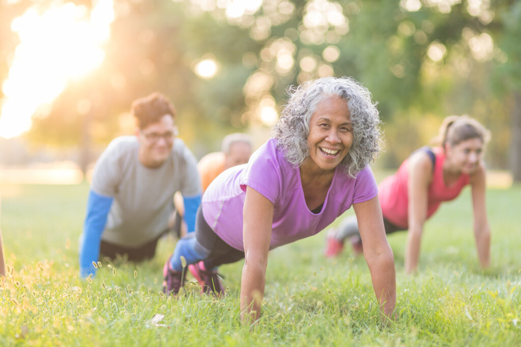 A multiethnic, coed group of various ages works out together outside in the early morning as the sun rises. They are doing pushups in the grass. The senior woman in front is looking at the camera and smiling.