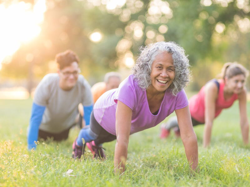 A multiethnic, coed group of various ages works out together outside in the early morning as the sun rises. They are doing pushups in the grass. The senior woman in front is looking at the camera and smiling.
